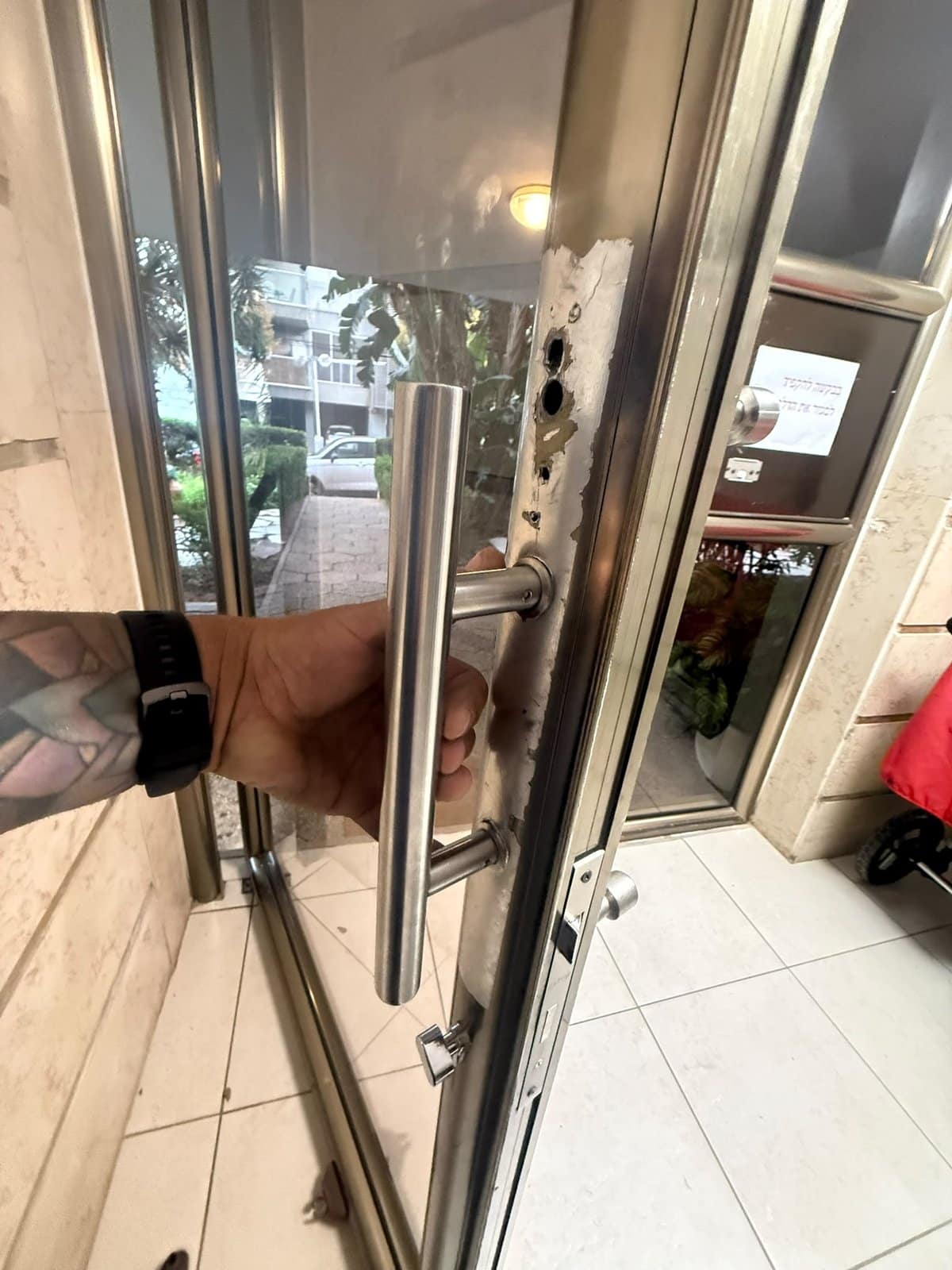 דלת זכוכית בכניסה לבניין ברמת גן לפני התיקון, עם חורי הברגה חשופים בפרופיל ליד הידית